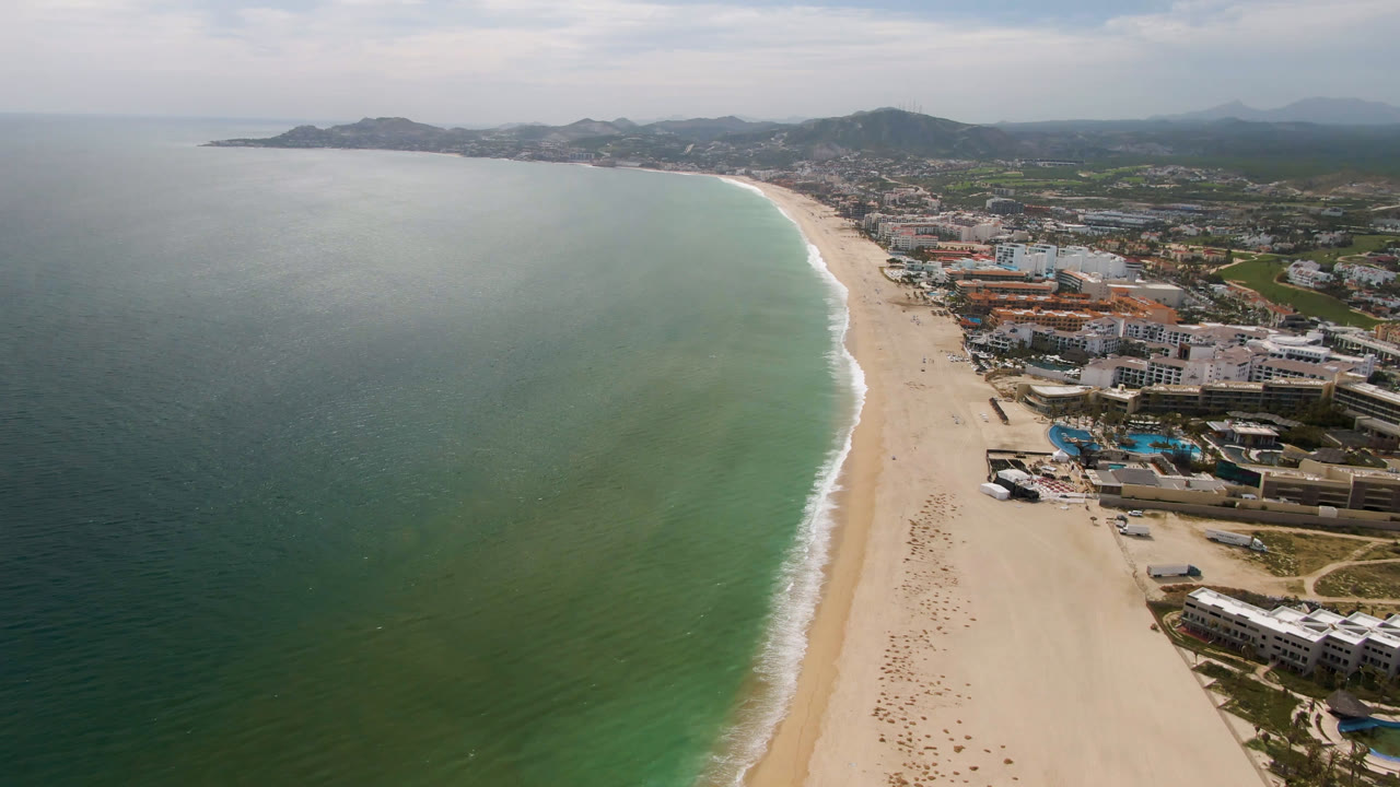 Wide aerial coastline with green water, pale sand, and a town behind the beach.