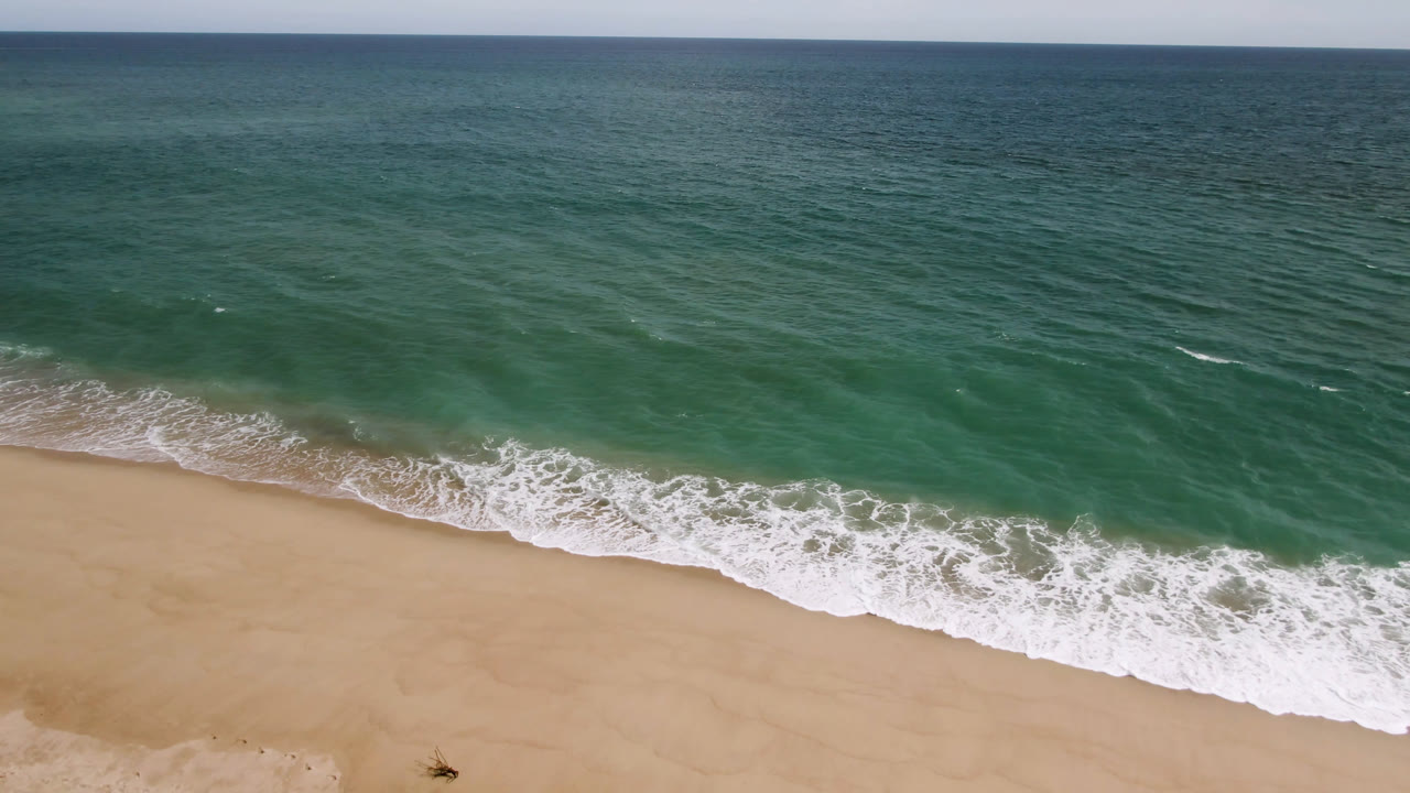 Aerial view in San Jose del Cabo, Mexico, with teal ocean filling the upper frame, white surf crossing the shore, sandy foreground, and no visible people.