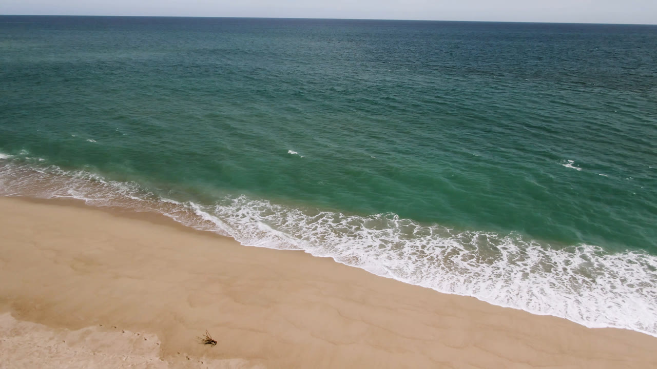 San Jose del Cabo aerial shoreline frame with a wider band of breaking foam over wet sand, open teal water above, and no visible people.