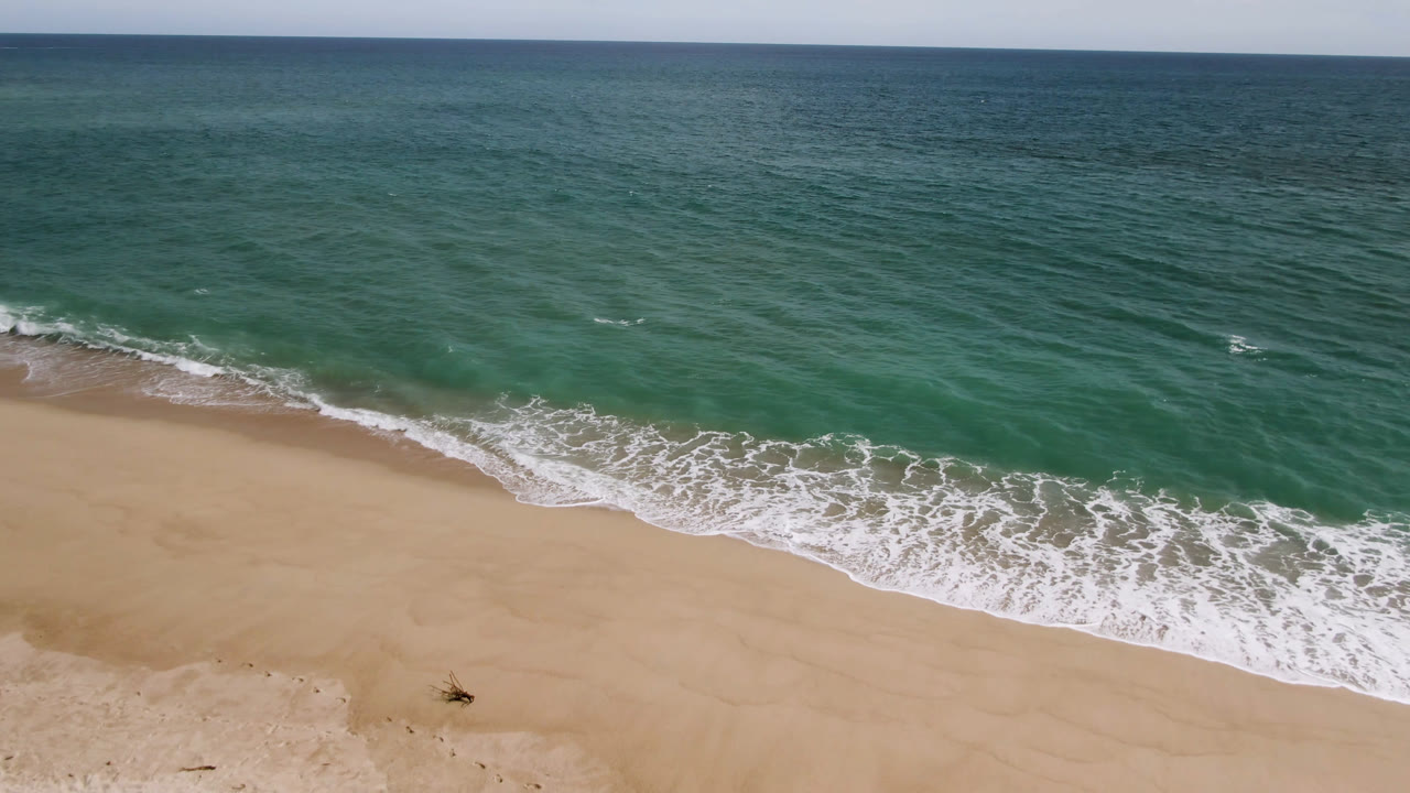 Aerial beach view with surf curling onto sand and a level ocean horizon.