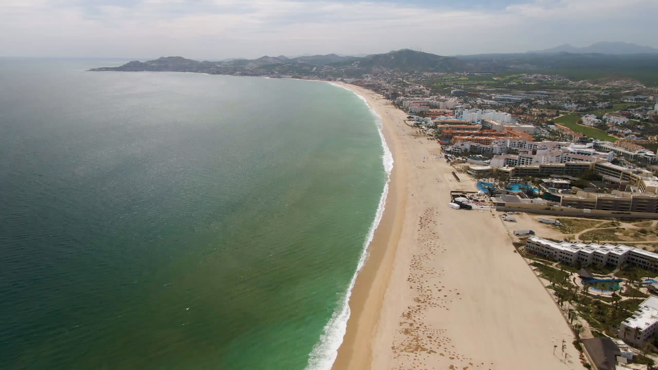 High aerial view over San Jose del Cabo with green-blue ocean, a pale curving beach, beachfront buildings, hazy hills, and no visible people.