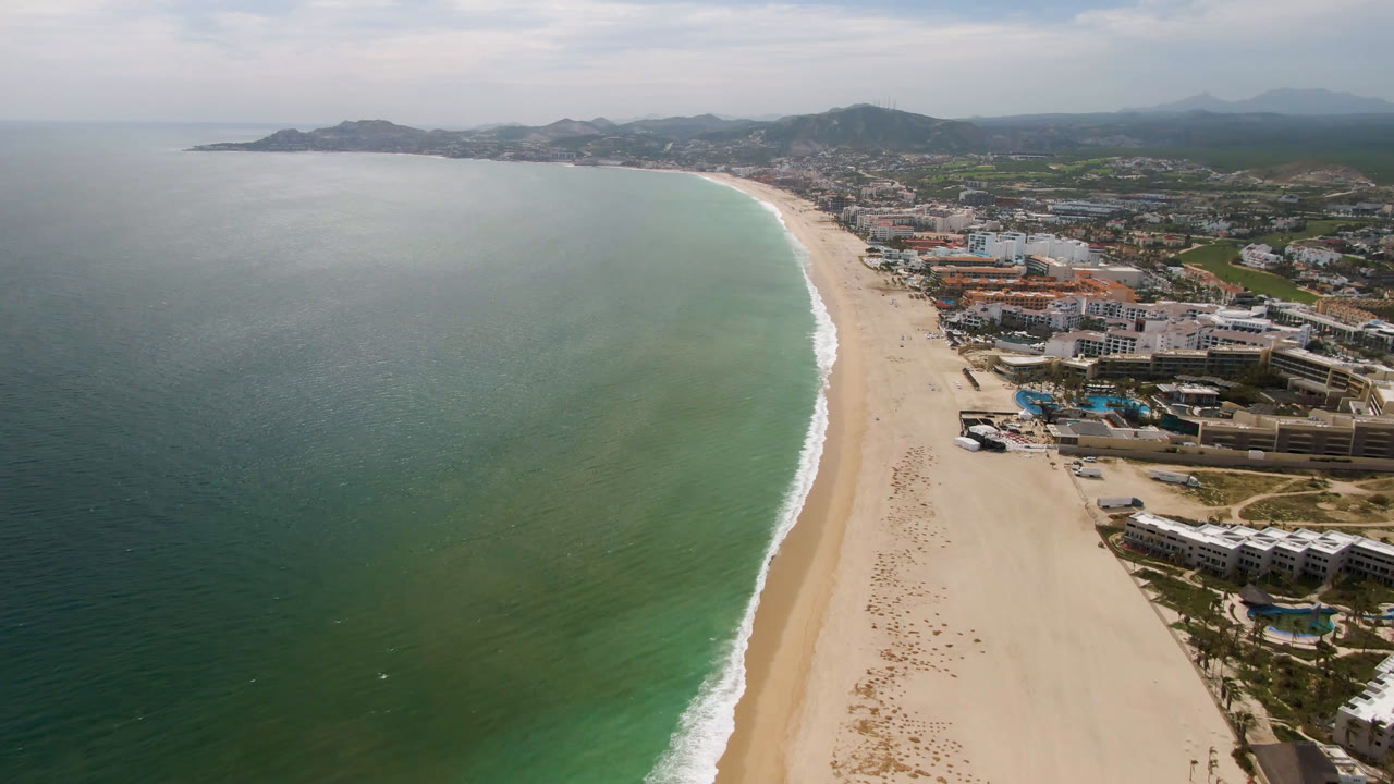 Drone-height view along San Jose del Cabo beach, with the ocean filling the left side, a long sand strip through the center, beachfront buildings to the right, and no visible people.