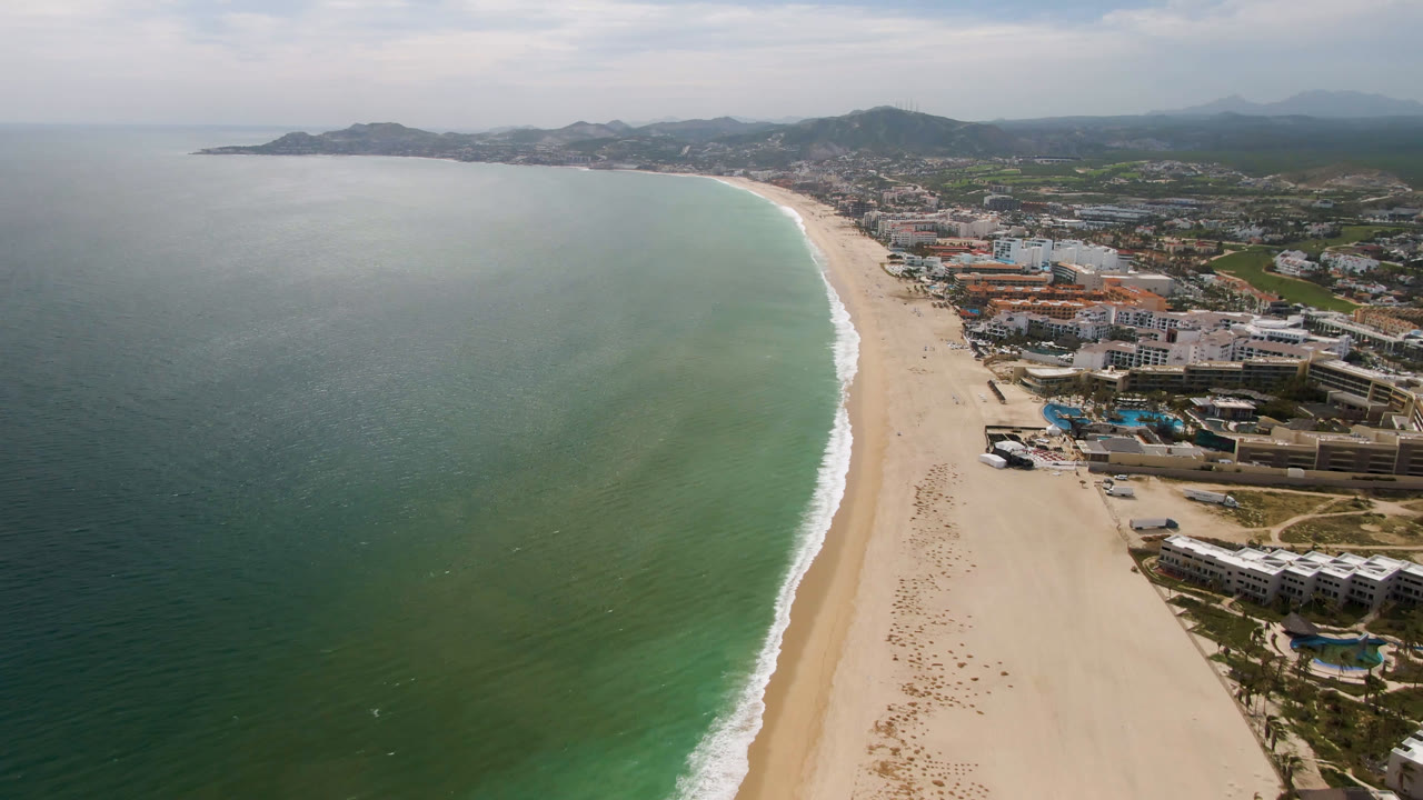 Aerial coastline view with dark green water, pale sand, surf, buildings, and hills.