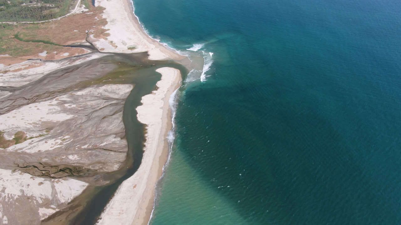 Opening aerial frame over San Jose del Cabo coastline, with a pale sand spit dividing dark inlet water from blue ocean and no visible people.