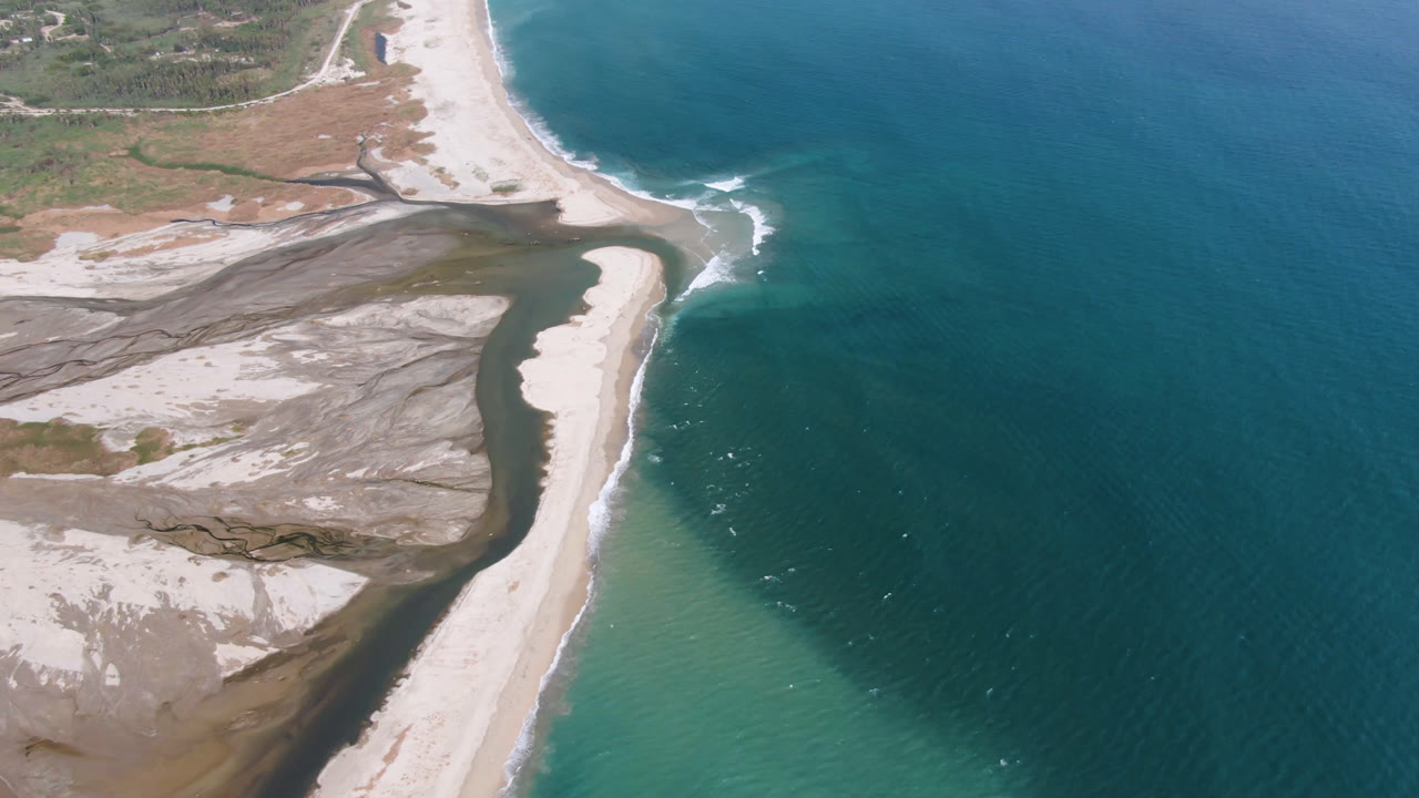Opening aerial frame over San Jose del Cabo showing pale sand banks, dark inlet water, blue ocean, white surf lines, and no visible people.