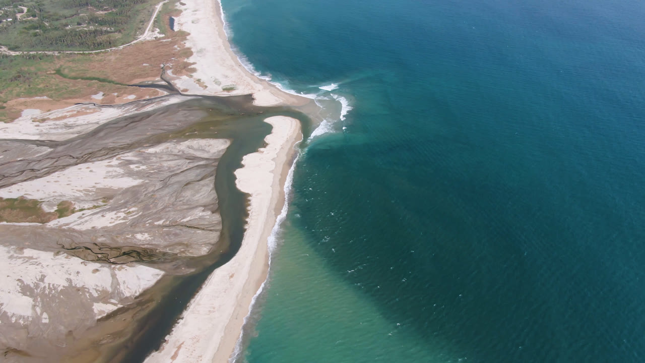 Aerial frame over San Jose del Cabo with the curved sand spit more centered between dark inland water and blue ocean, with no visible people.