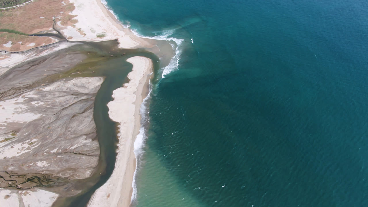 Aerial frame over San Jose del Cabo showing a pale sand spit between teal ocean water and a darker inlet, with exposed flats on the left and no visible people.