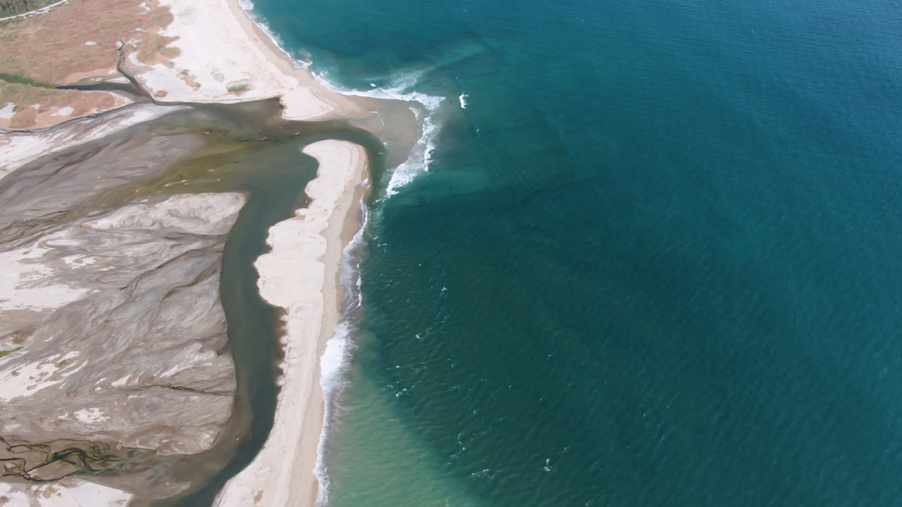 Overhead San Jose del Cabo coastline frame with white surf curling beside the narrow sand bank and darker shallow water running along the inland side, with no visible people.