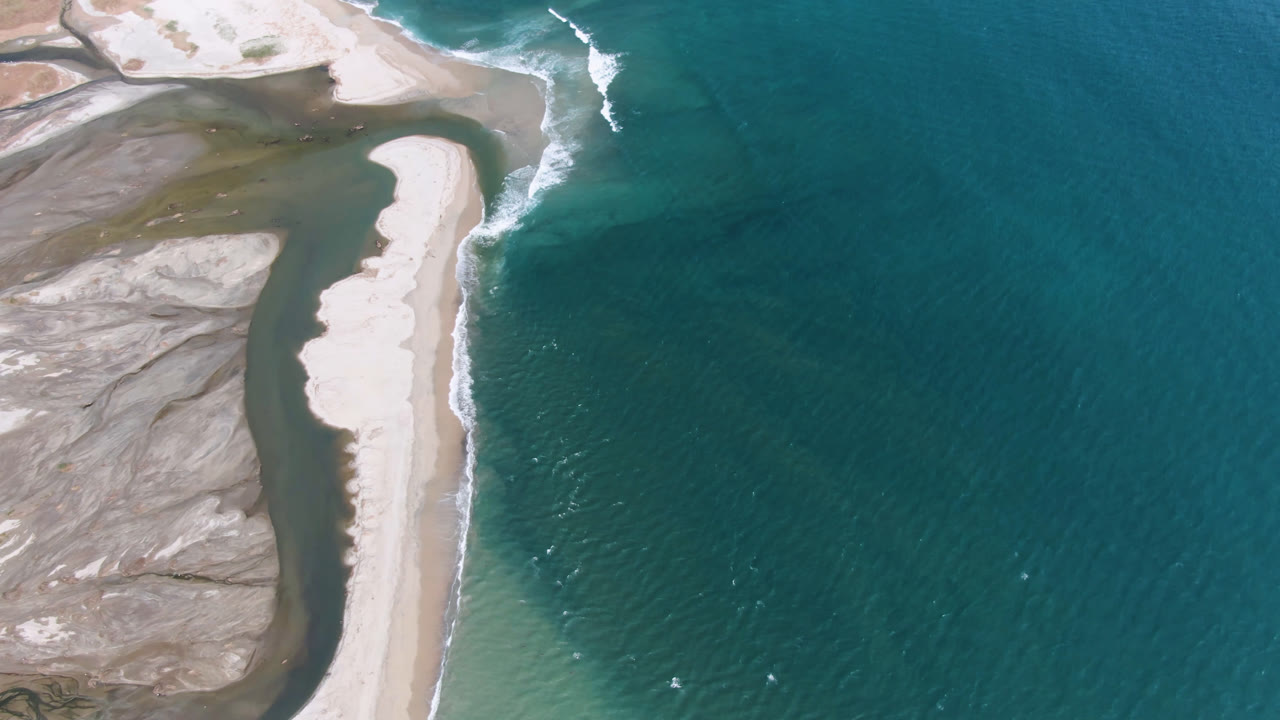 A high aerial frame over San Jose del Cabo shows a pale sandbar curving between shallow lagoon water and darker blue ocean, with no visible people.