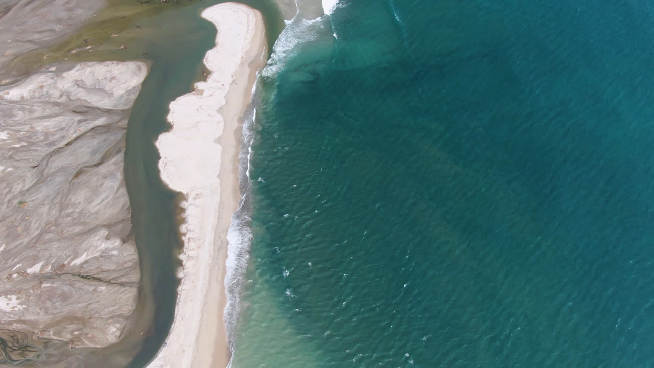 Overhead aerial frame in San Jose del Cabo showing a pale sand bar between teal ocean and a darker channel, with rocky flats at left and no visible people.