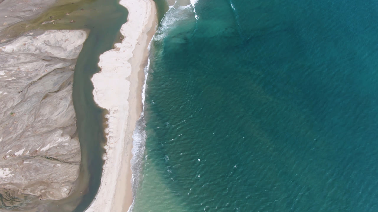 Aerial view in San Jose del Cabo with the long sand bank centered, white wave foam on the ocean side, muted flats to the left, and no visible people.