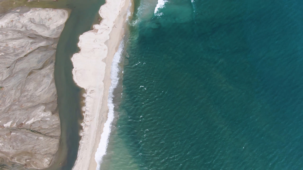 Overhead aerial frame from San Jose del Cabo showing a pale beach between teal ocean and gray rocky terrain, with a narrow inland water channel and no visible people.