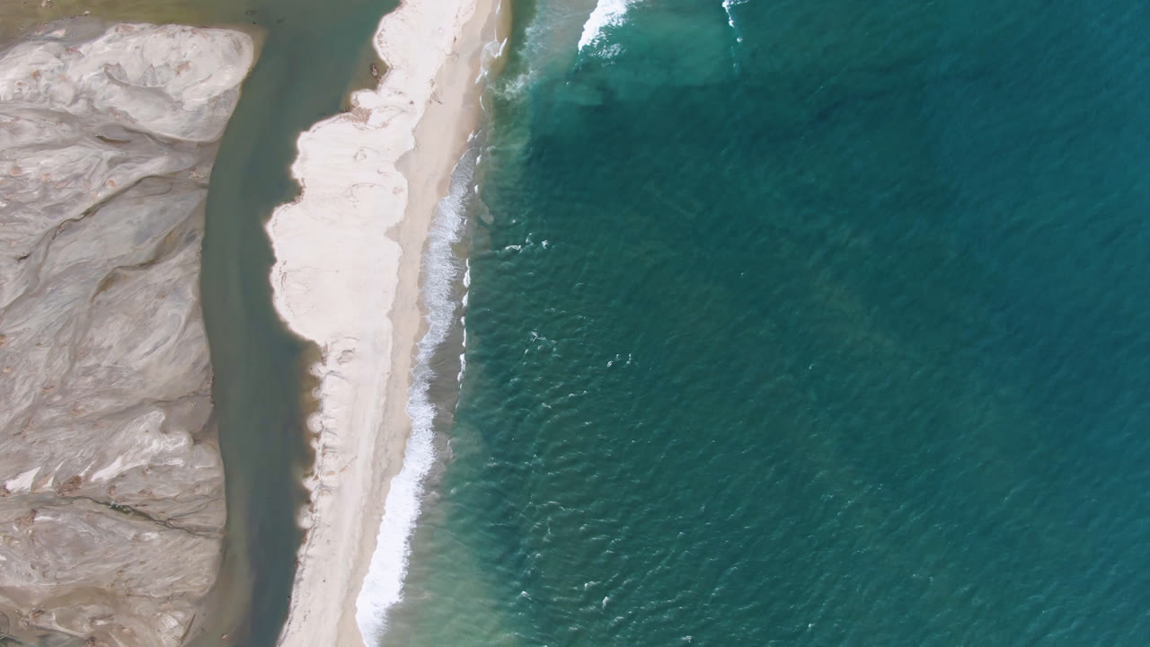 A San Jose del Cabo aerial still shows white surf edging a pale sand strip, with teal ocean filling the right side and rocky land along the left, with no visible people.