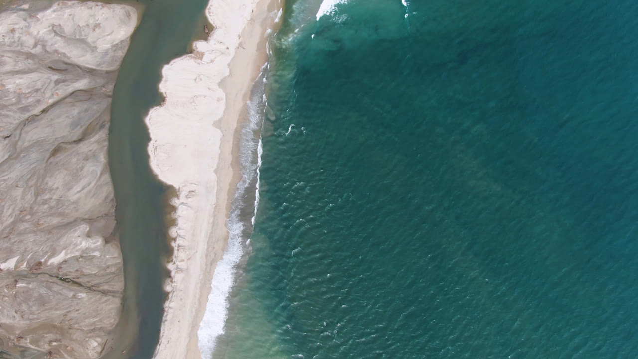 Top-down coastal view with beach, breaking waves, rocky terrain, and open ocean.