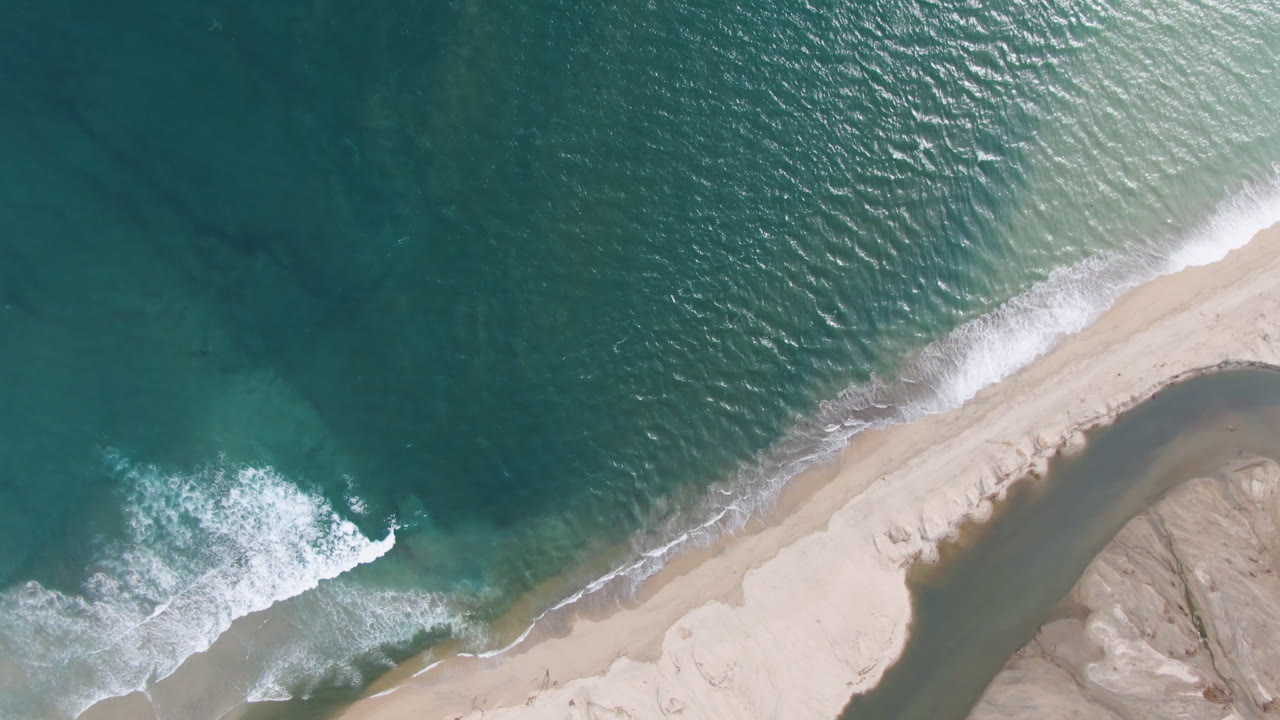 Top-down aerial view in San Jose del Cabo shows teal water, a diagonal strip of pale beach, white surf breaking at the lower left, and no visible people.
