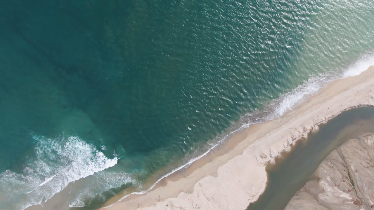 Aerial frame from San Jose del Cabo shows rippled teal ocean across the top, a diagonal sandy shoreline, a dark water channel near the lower right, and no visible people.