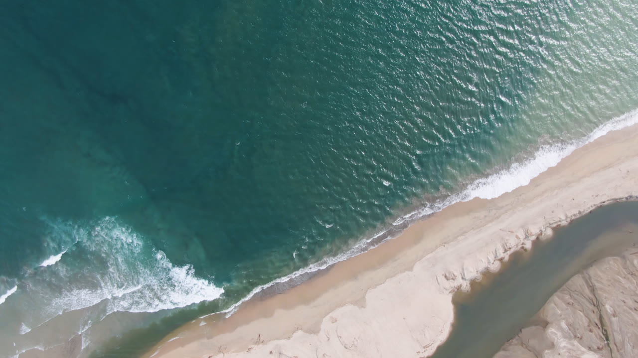 Overhead view of the San Jose del Cabo shoreline with teal water above a diagonal strip of sand, white surf at left, a dark tidal channel at lower right, and no visible people.