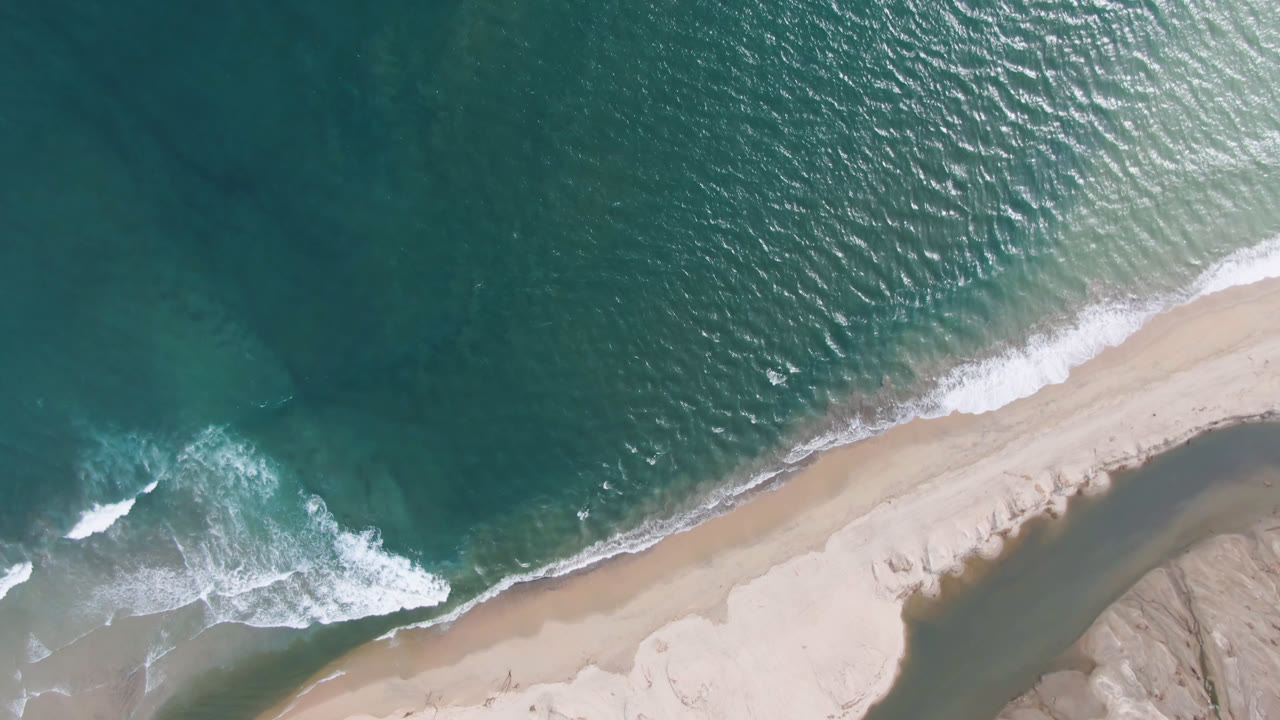 Aerial frame from San Jose del Cabo showing rippled teal ocean across the top, a pale diagonal beach, broken white foam at left, and no visible people.