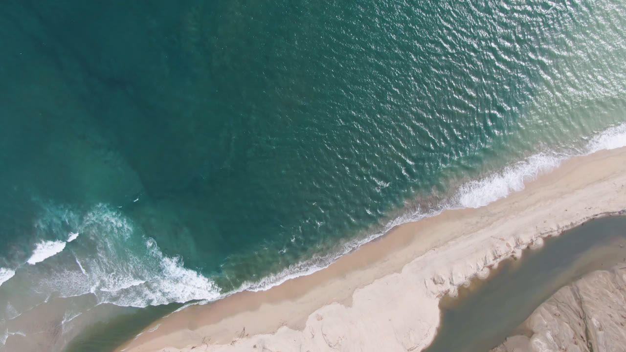 Drone view of ocean meeting a sandy spit with surf and shallow water.