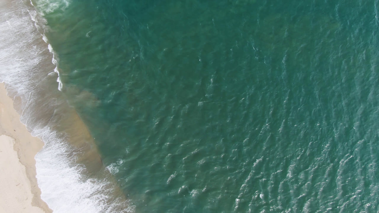 Overhead view of turquoise water and a sandy San Jose del Cabo shoreline entering from the left, with white surf at the beach edge and no visible people.
