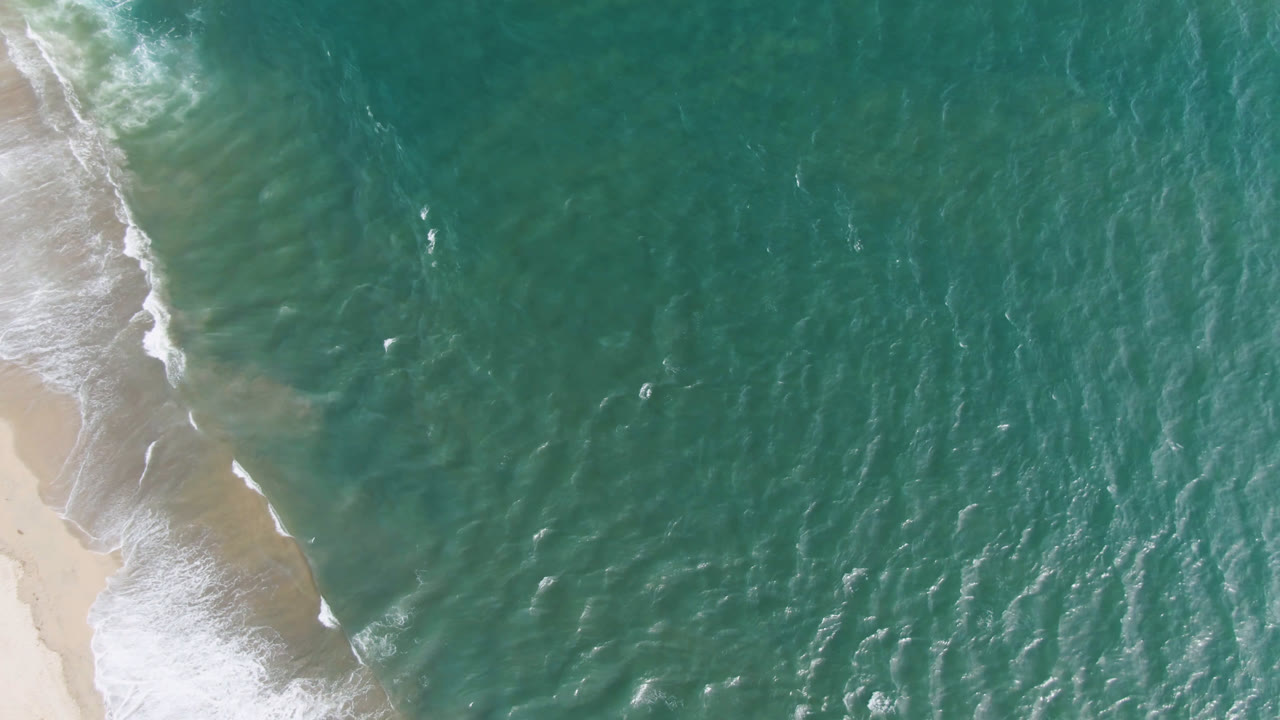 Aerial frame over San Jose del Cabo water, with pale sand along the left border, foamy shore break, and broad teal ocean showing no visible people.