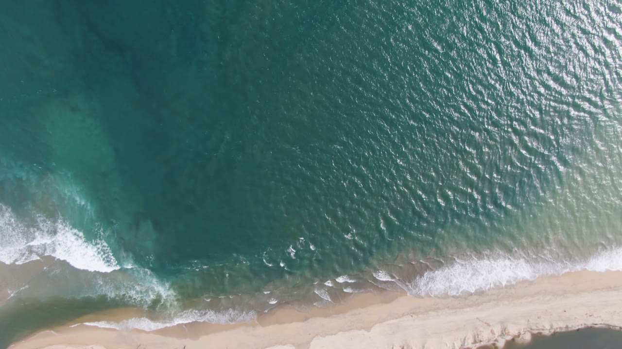 High overhead view of teal water meeting pale sand in San Jose del Cabo, with white surf crossing the lower frame and no visible people.