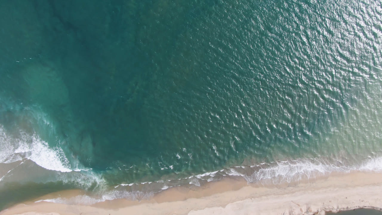 Aerial frame over San Jose del Cabo shows green-blue water covering most of the view, surf curling toward the sand, and no visible people.
