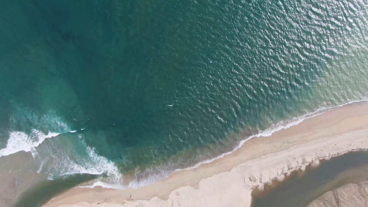 Overhead view of teal water and white surf reaching a pale beach in San Jose del Cabo, Mexico, with no visible people.