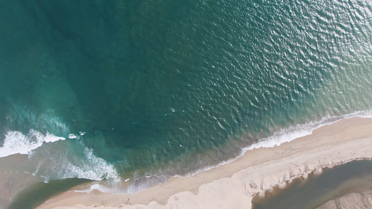 Top-down San Jose del Cabo beach frame with white foam tracing a diagonal shoreline, teal water above, pale sand below, and no visible people.