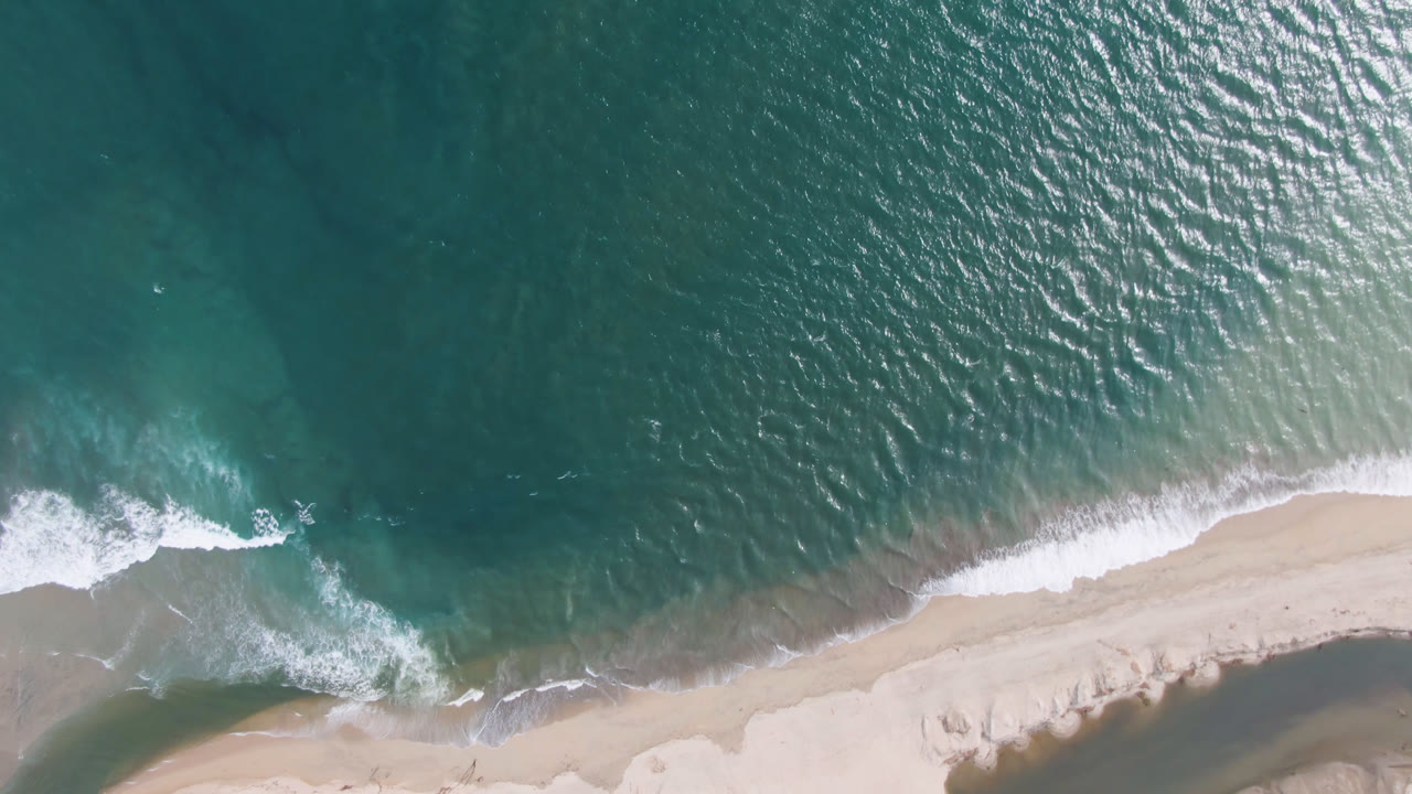 Drone view of ocean surface, surf, and light sand.