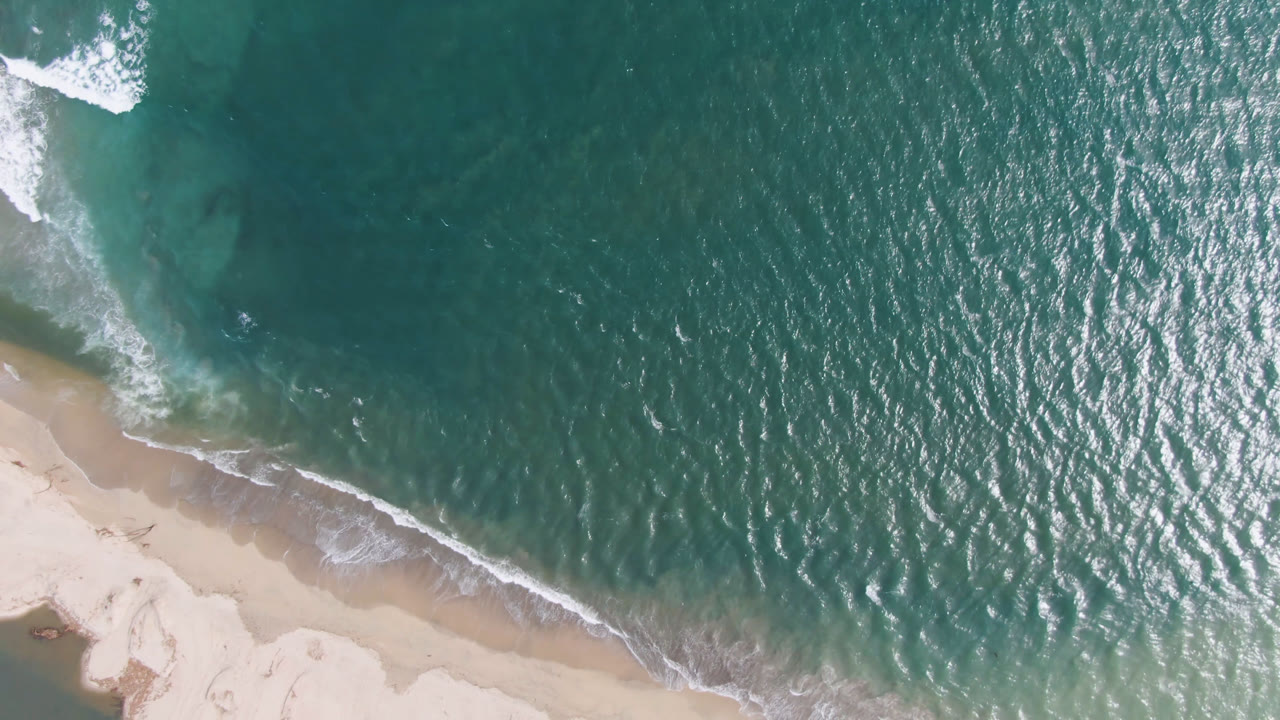 Overhead view of teal water and pale beach in San Jose del Cabo, with white surf entering from the upper left and no visible people.