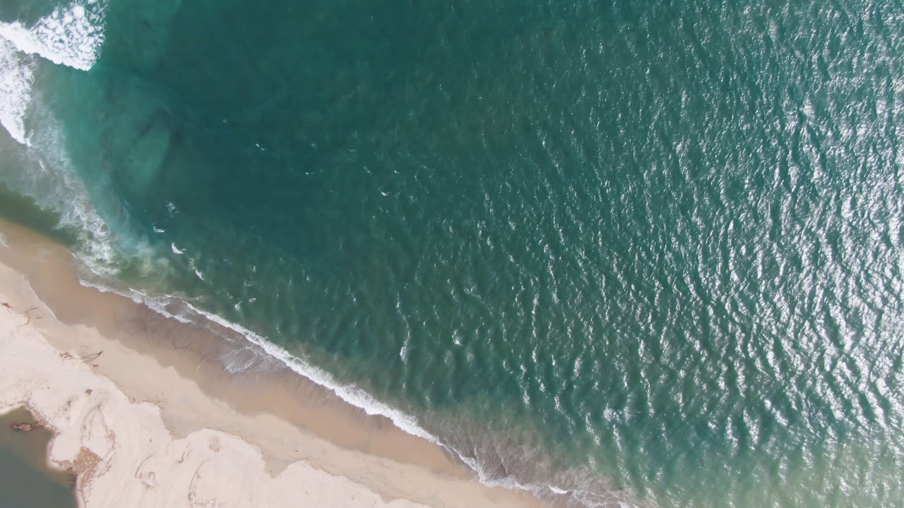 Aerial frame over San Jose del Cabo shoreline, showing rippled teal water, a diagonal strip of sand, and no visible people.