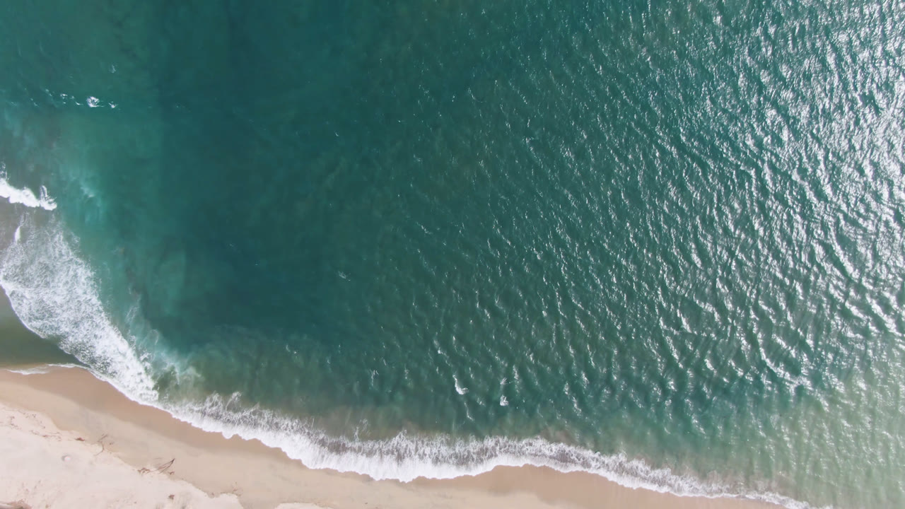 Overhead view of teal water and white surf reaching pale sand in San Jose del Cabo, Mexico, with no visible people.