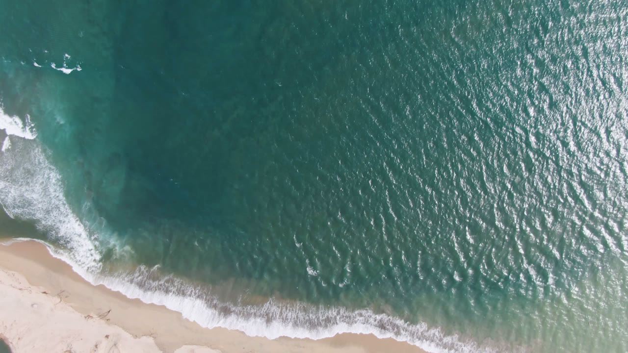 Sunlit ripples spread across teal ocean above a narrow strip of sand in San Jose del Cabo, Mexico, with no visible people.