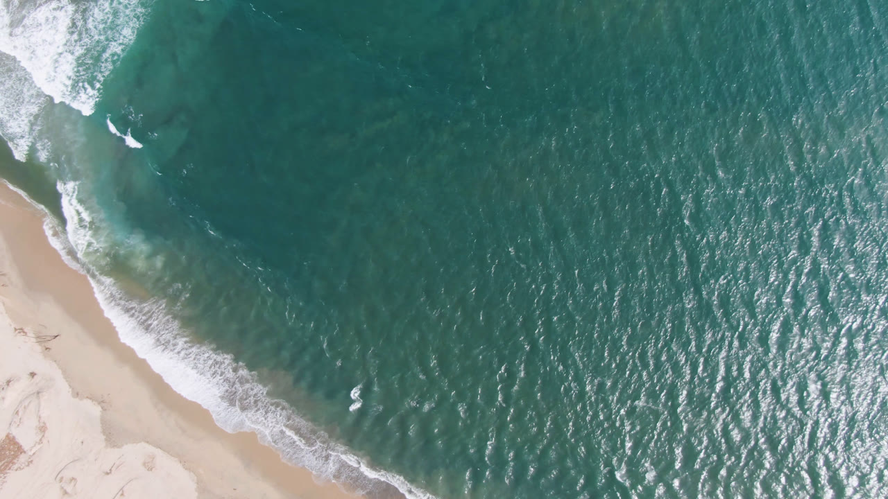 A high aerial view over San Jose del Cabo shows teal water and white surf crossing toward pale sand, with no visible people.