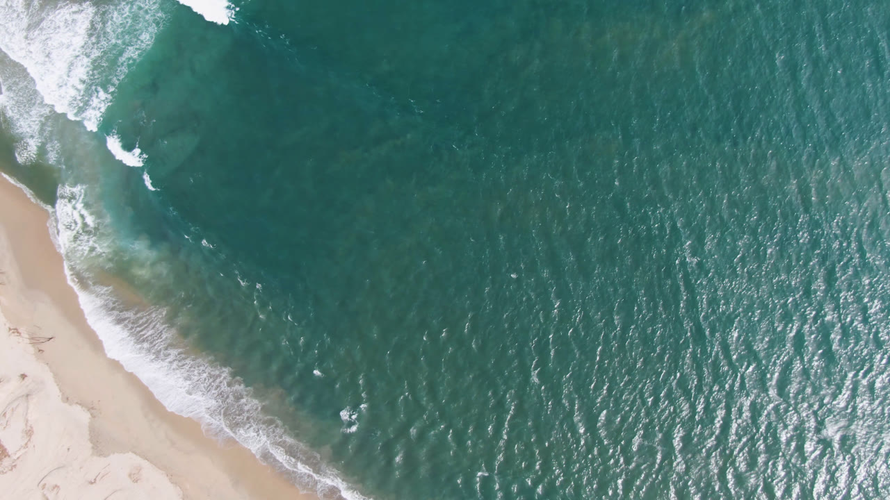 The San Jose del Cabo shoreline appears from above with a wider band of pale sand on the right and foam curling along the waterline, with no visible people.