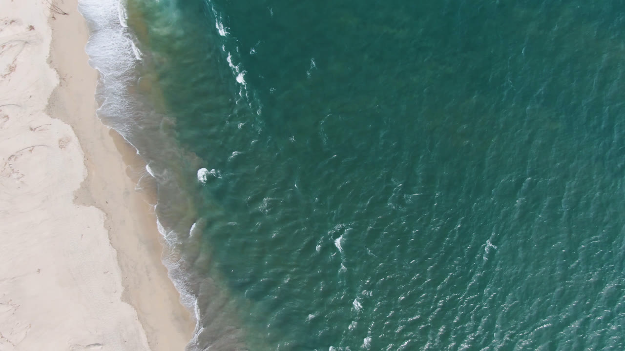 Top-down aerial frame from San Jose del Cabo showing teal ocean along a pale sand edge with small white wave breaks and no visible people.