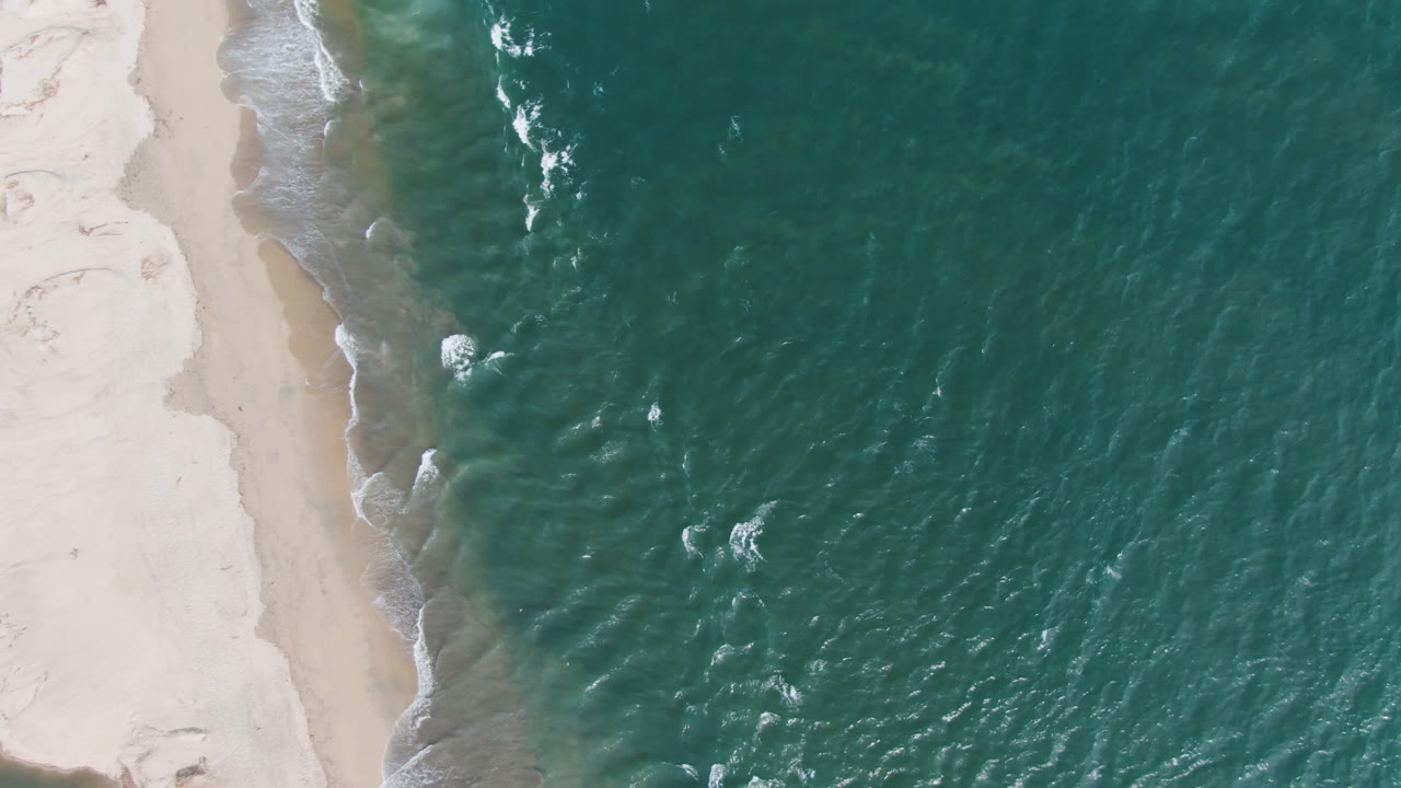 Overhead San Jose del Cabo shoreline frame with textured green-blue water pressing against a narrow white surf line and no visible people.