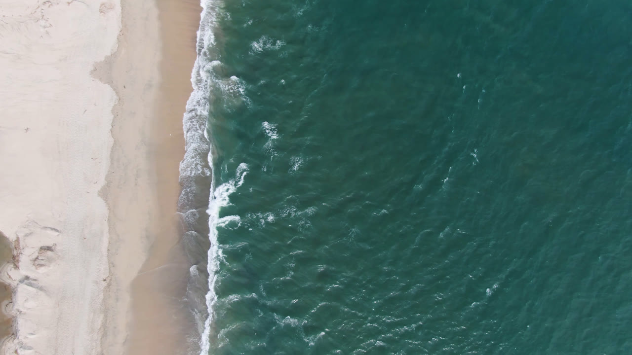 Top-down aerial frame from San Jose del Cabo showing teal ocean pressed against pale sand, with white foam along the shore and no visible people.