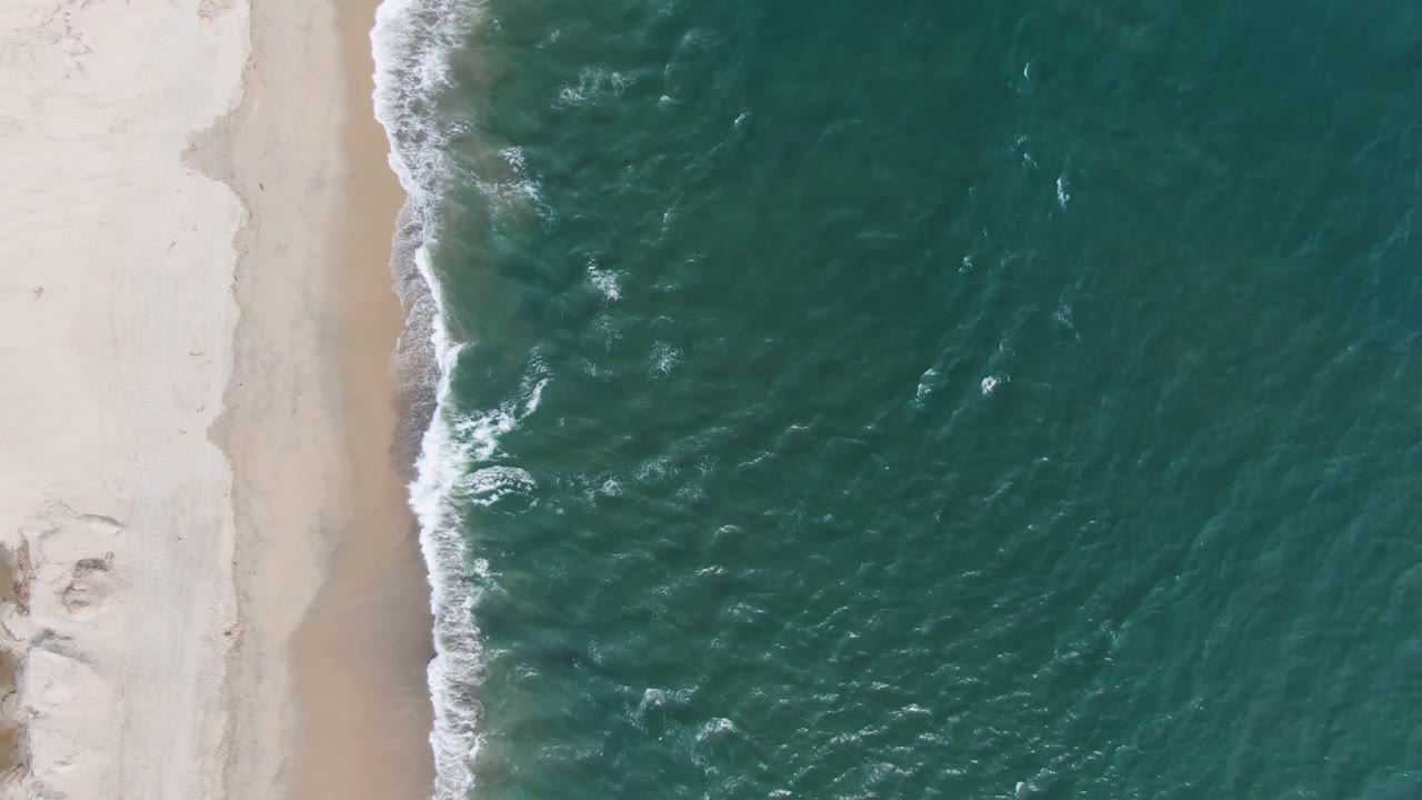 San Jose del Cabo aerial frame with textured green-blue water filling most of the view, a narrow band of white surf, pale sand, and no visible people.