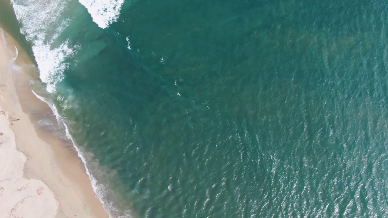 Aerial view of San Jose del Cabo shoreline with pale sand on the left, teal water across the frame, a white wave breaking near the top, and no visible people.