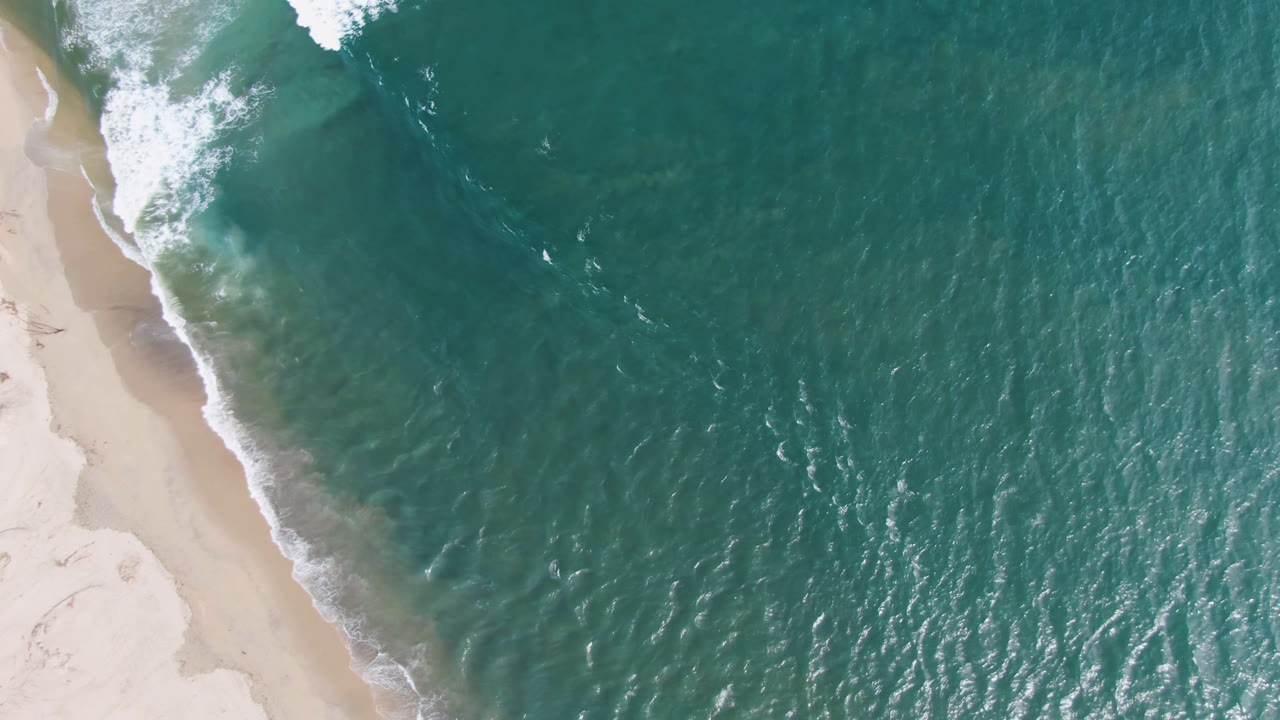 Aerial frame over San Jose del Cabo beach showing the sandy shoreline running diagonally down the left side, rippled teal ocean to the right, and no visible people.