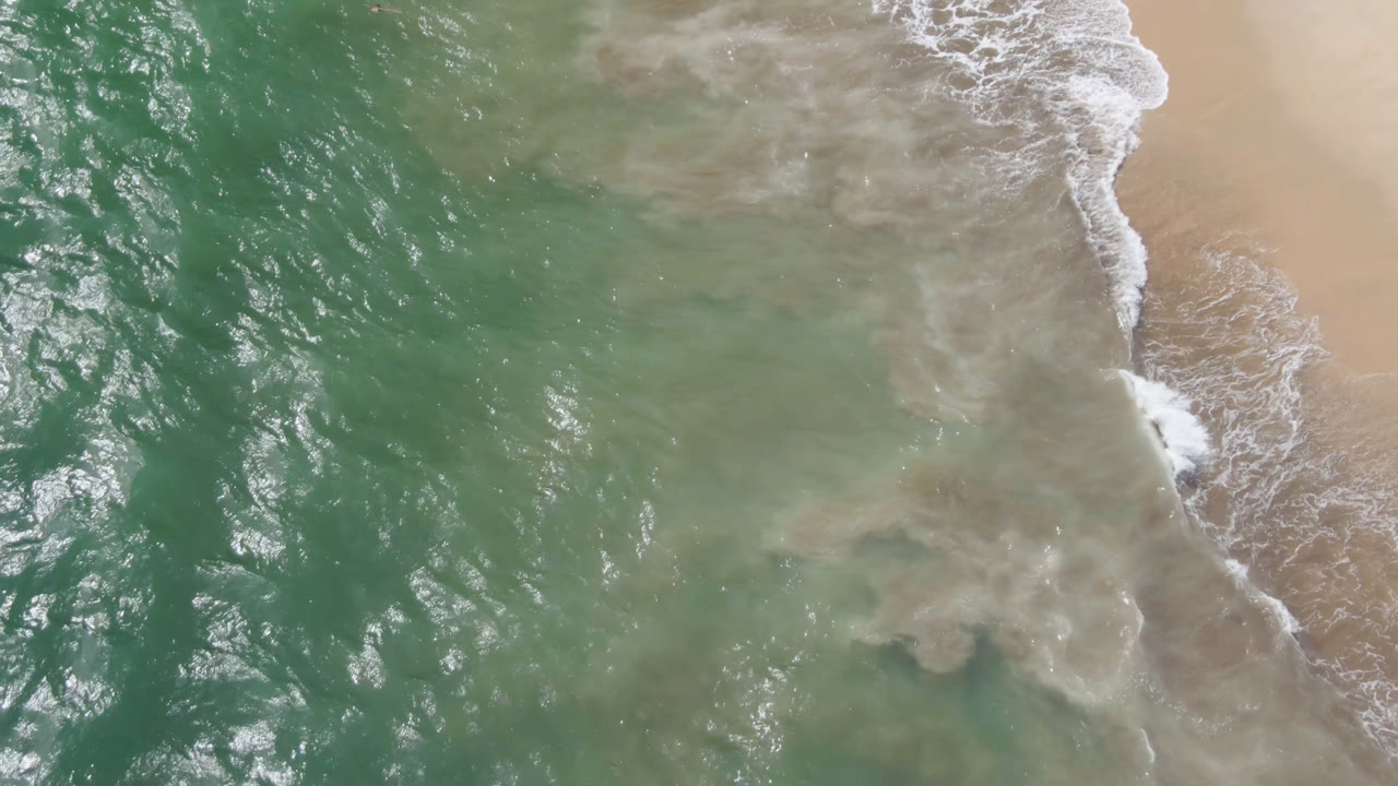 Aerial frame from San Jose del Cabo showing green ocean water across the left and white surf curling onto tan beach at right, with no visible people.