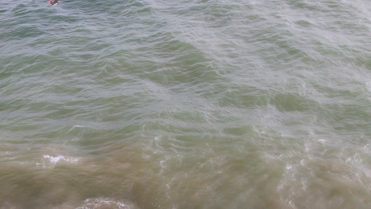 Overhead view of green-blue surf water in San Jose del Cabo with pale foam lines and yellowish sand tones beneath the surface, with no recognizable people visible.