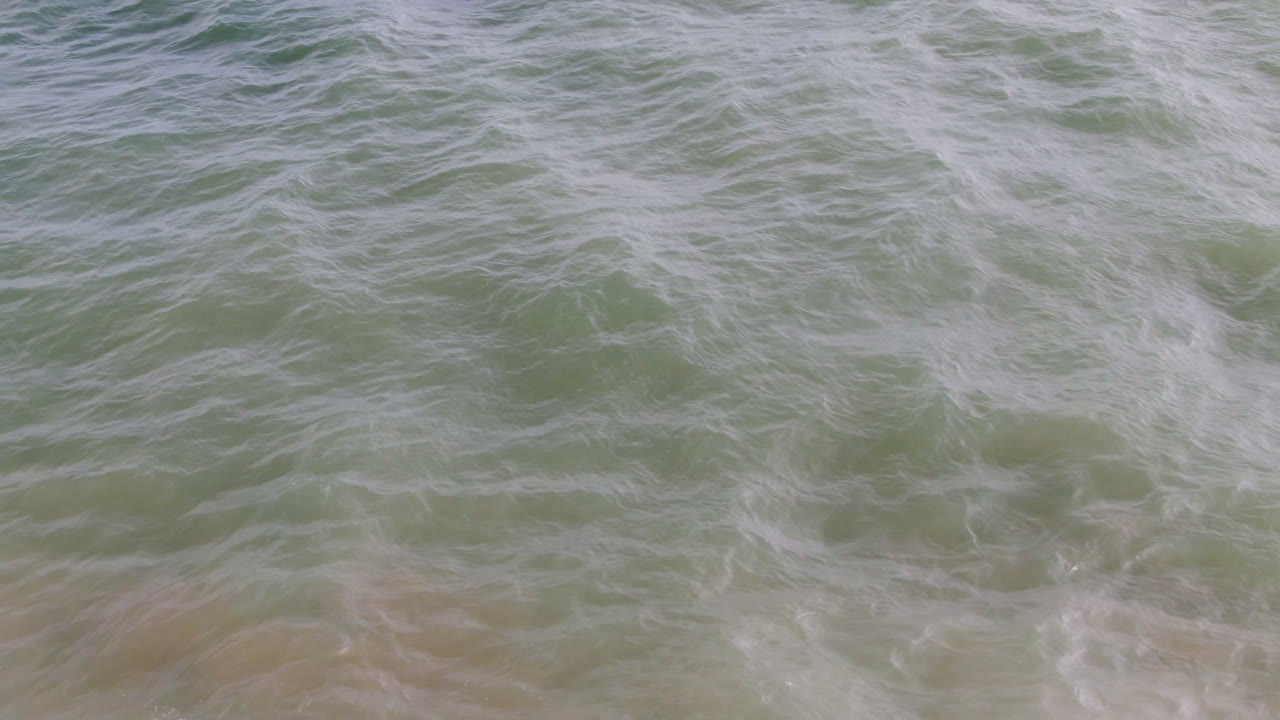 Aerial frame of hazy San Jose del Cabo ocean water with broken white foam patterns spreading across green and yellow shallow water, with no recognizable people visible.