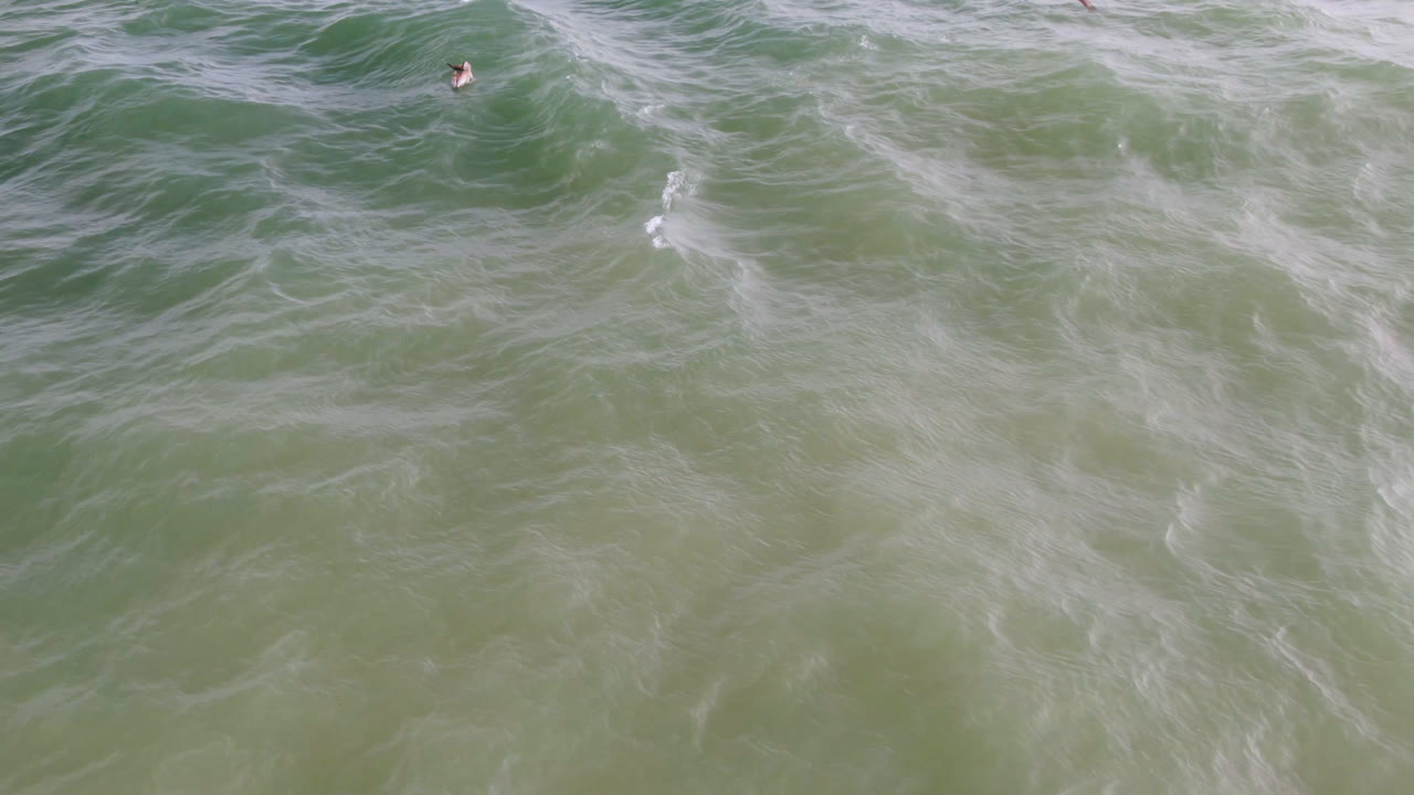 Overhead green ocean waves with distant surfers near the top of frame.