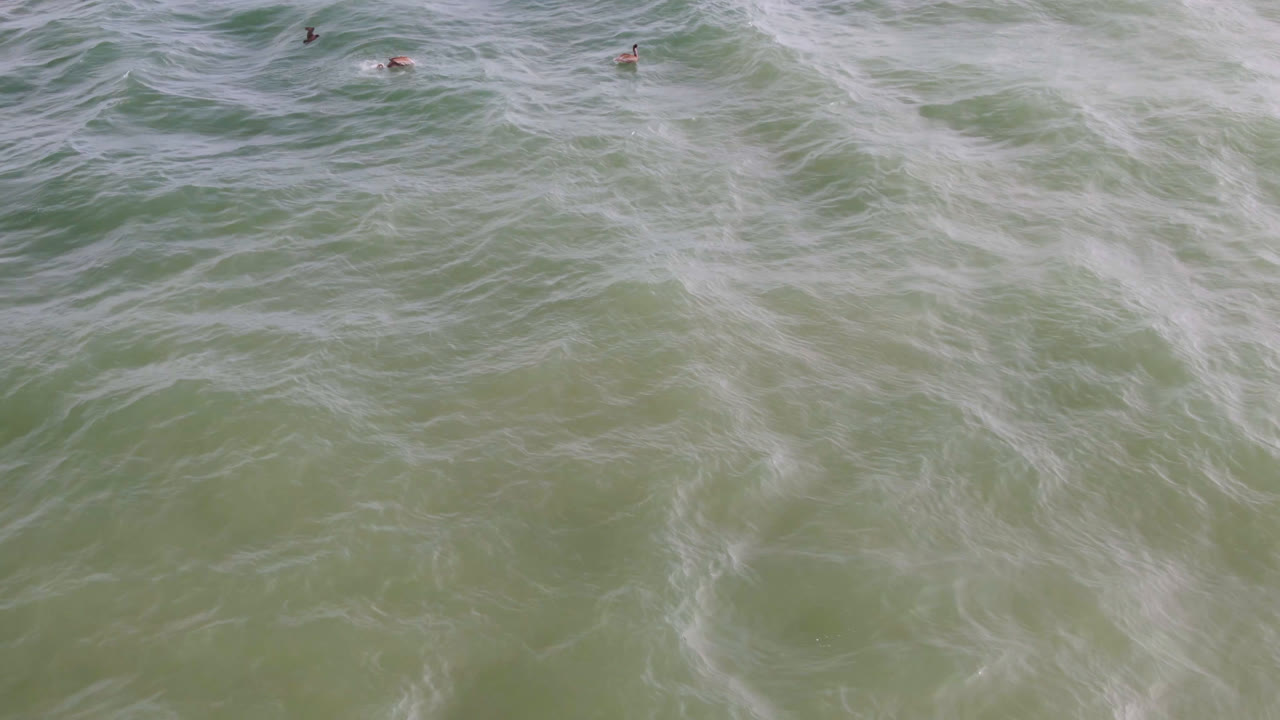 Top-down ocean water with rolling foam texture and a tiny surfer near the upper right.