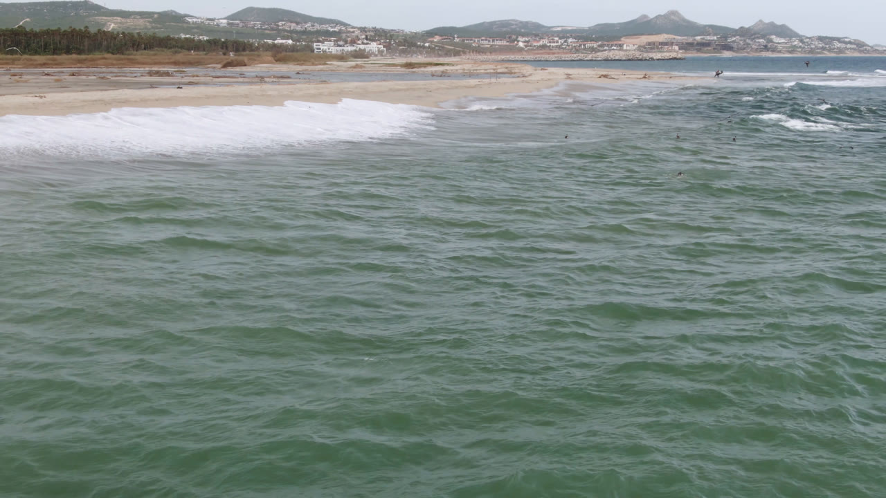 Aerial view over green ocean toward a sandy beach with small waves and distant surfers.