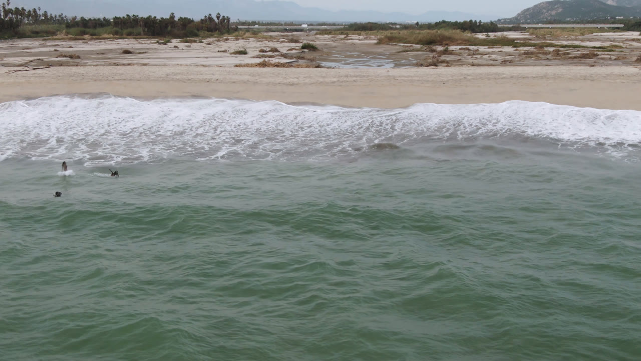 San Jose del Cabo beach seen from above, with a wide white surf band crossing the frame and several small surfers floating beyond the shore break.