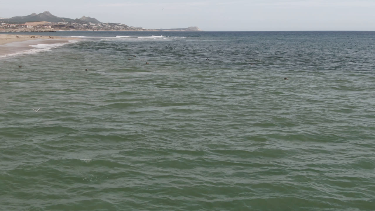 Opening frame over green-blue ocean at San Jose del Cabo, with sandy beach and low hills in the distance and no visible people.